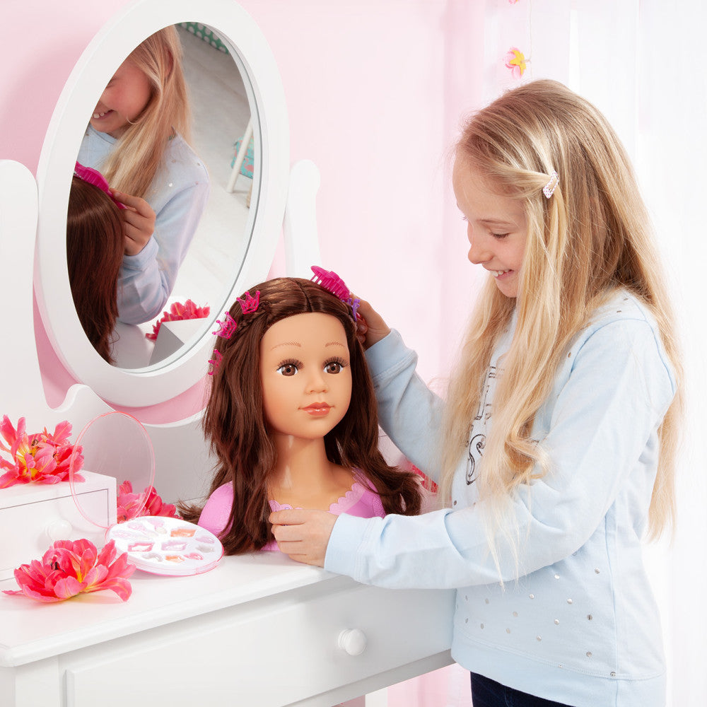 Child modeled by a girl styling the hair of a brunette doll head with brown hair at a white vanity mirror with pink flowers