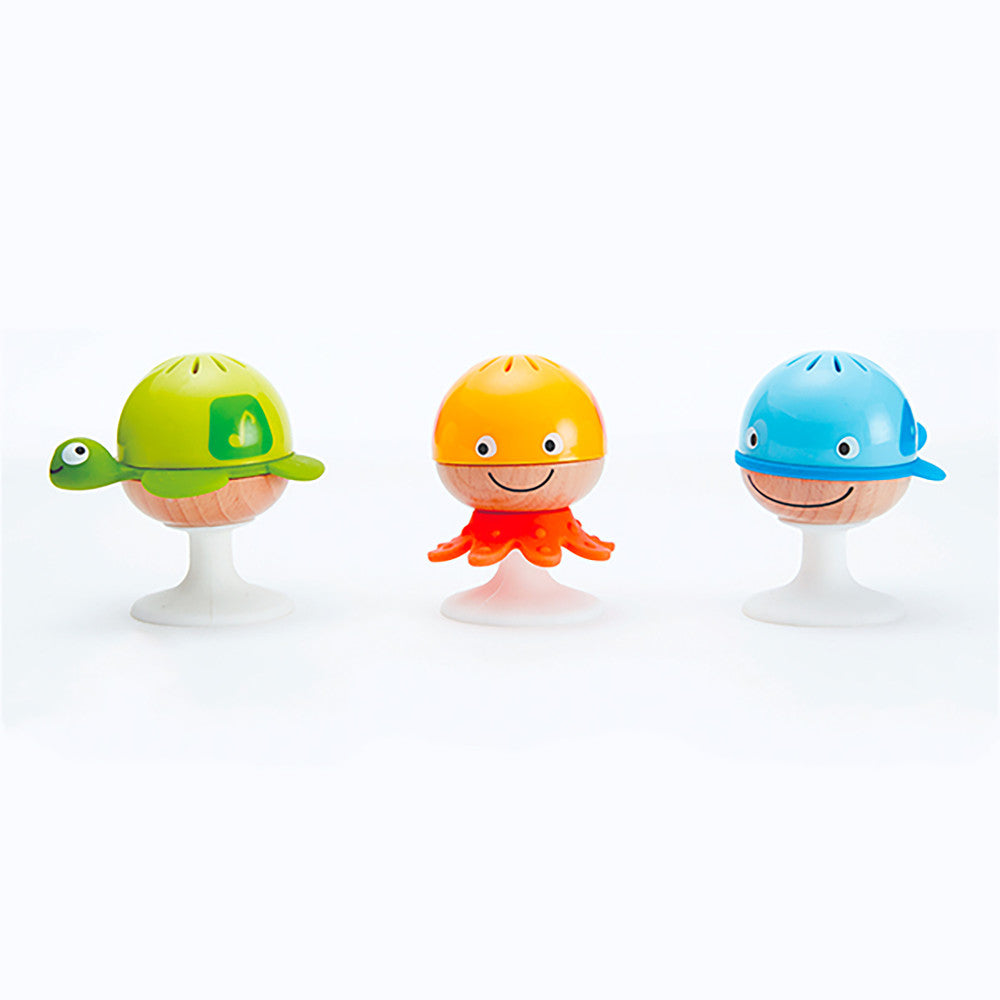 Three colorful suction-cup rattles with smiling faces: green turtle, yellow octopus, and blue whale, each with a wooden ball body and white suction-cup base