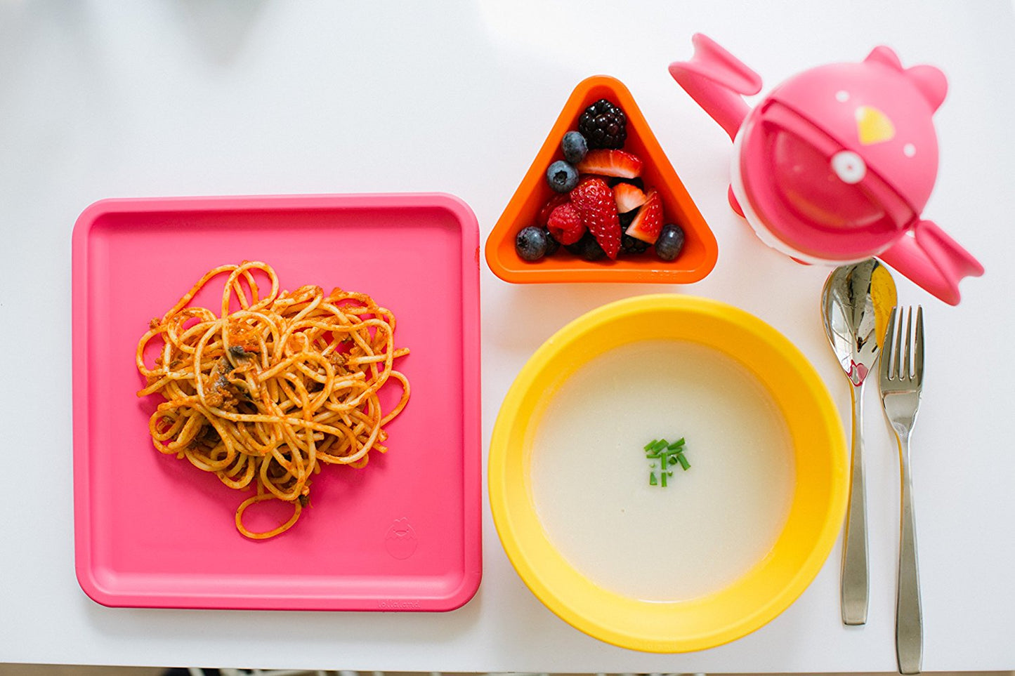 Colorful kid-friendly dining set with pink square plate holding pretzel snacks, yellow bowl with soup and green garnish, orange triangle bowl with berries, pink