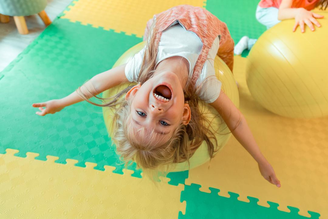 Child with headphones lying upside down on colorful foam puzzle mat, laughing with arms spread