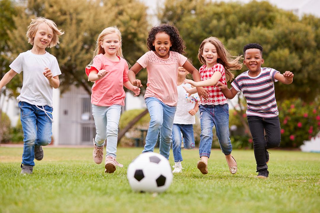 Five children running together on grass toward a soccer ball, playing outdoors in a sunny backyard with trees and a house in background
