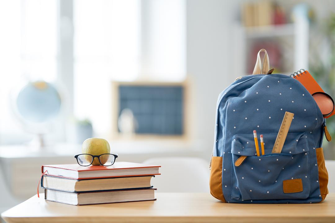 Back-to-school desk setup with stacked books, glasses, green apple, blue polka-dot backpack with tan accents, pencils, and headphones on wooden table in bright