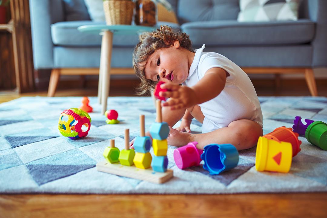 Toddler on model playing with colorful wooden stacking toys and shape sorters on a blue play mat in a living room