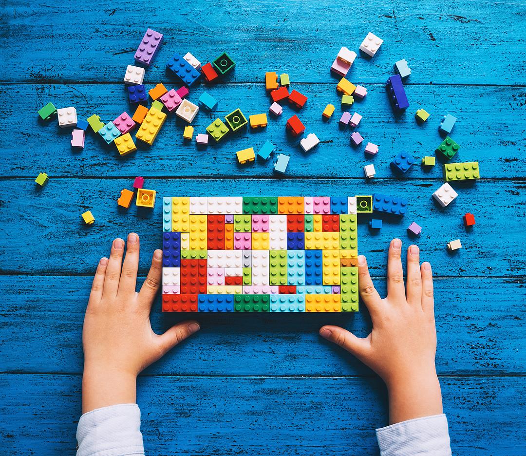 Child's hands assembling colorful building bricks on blue wooden surface with scattered bricks nearby, modeled by a child