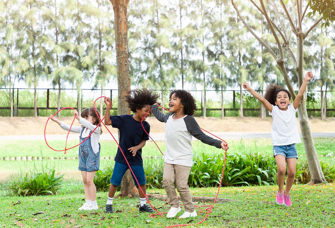 Four children playing outdoors in a park with red jump ropes, laughing and celebrating together on grass with trees in background