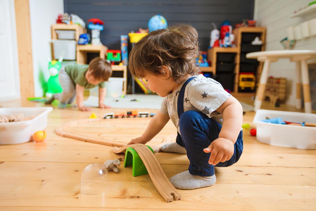 Two young children playing with wooden building blocks and track sets on a playroom floor surrounded by toys and storage shelves
