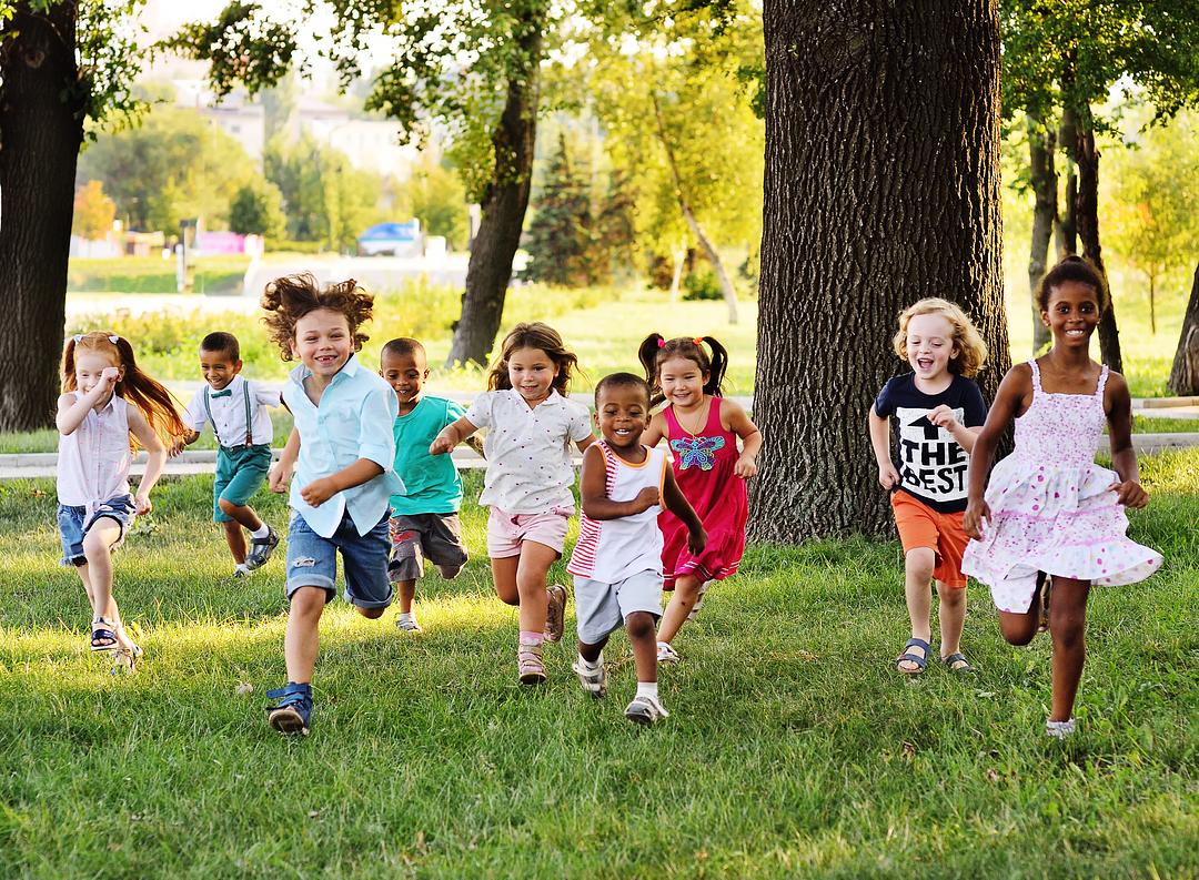 Group of diverse children running together outdoors on grass beneath large trees on a sunny day