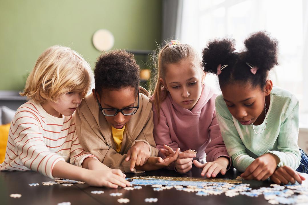 Four children working on a puzzle together at a table, modeled by children of diverse backgrounds wearing colorful clothing