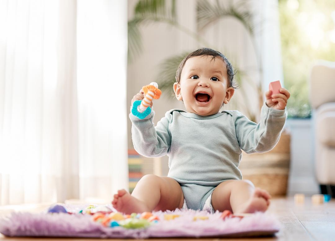 Toddler on model sitting on purple play mat, holding colorful teething ring and toy, smiling in bright living room