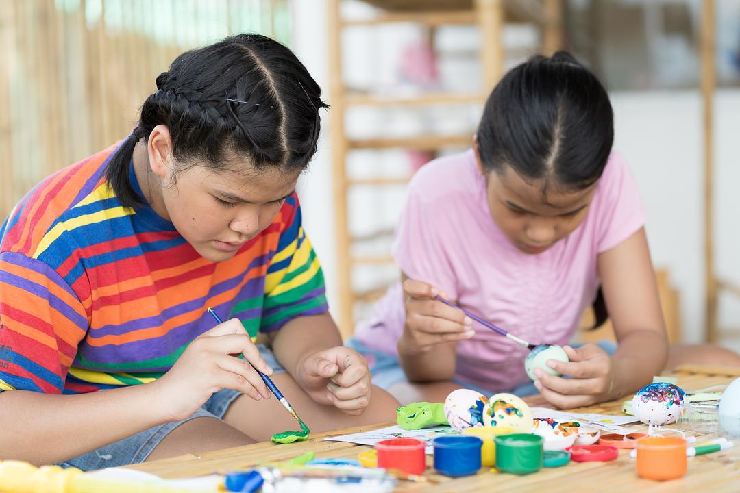 Two children painting crafts at a wooden table, modeled by children in colorful striped and pink shirts with paintbrushes and colorful paint containers