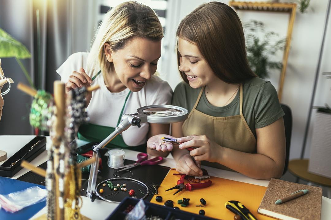 Adult and child modeled by a girl working together on a jewelry-making or crafting project at a table with a magnifying lamp, beads, tools, and supplies