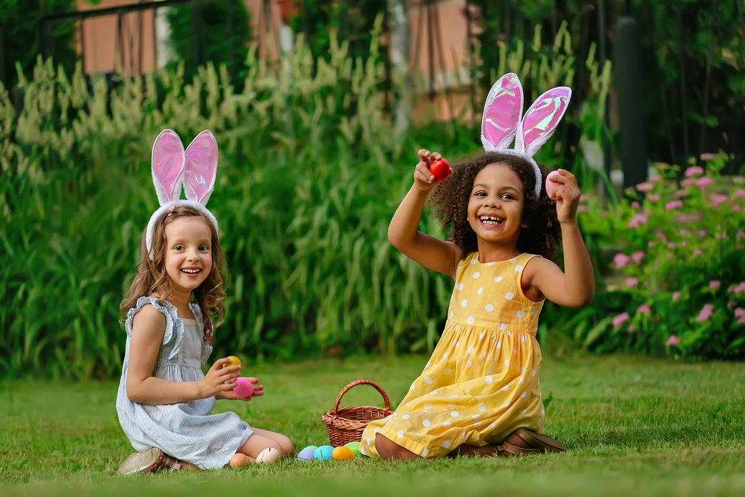Two children wearing bunny ear headbands sit on grass in a garden, holding Easter eggs and a wicker basket filled with colorful eggs