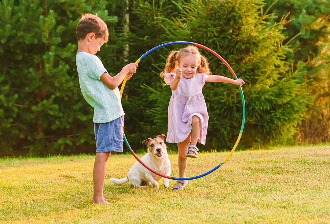 Two children and a small dog playing with a colorful hula hoop on a grass lawn with evergreen trees in the background