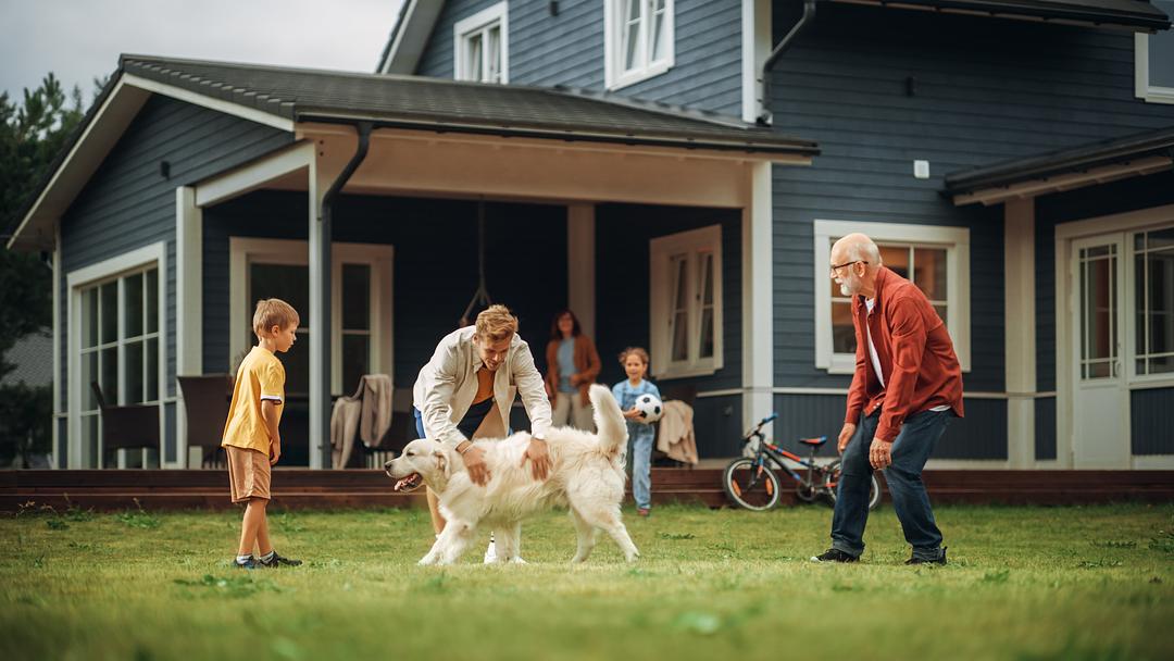 Multi-generational family playing together outdoors on grass in front of a modern blue and tan house, with children, adults, and a dog engaged in active play