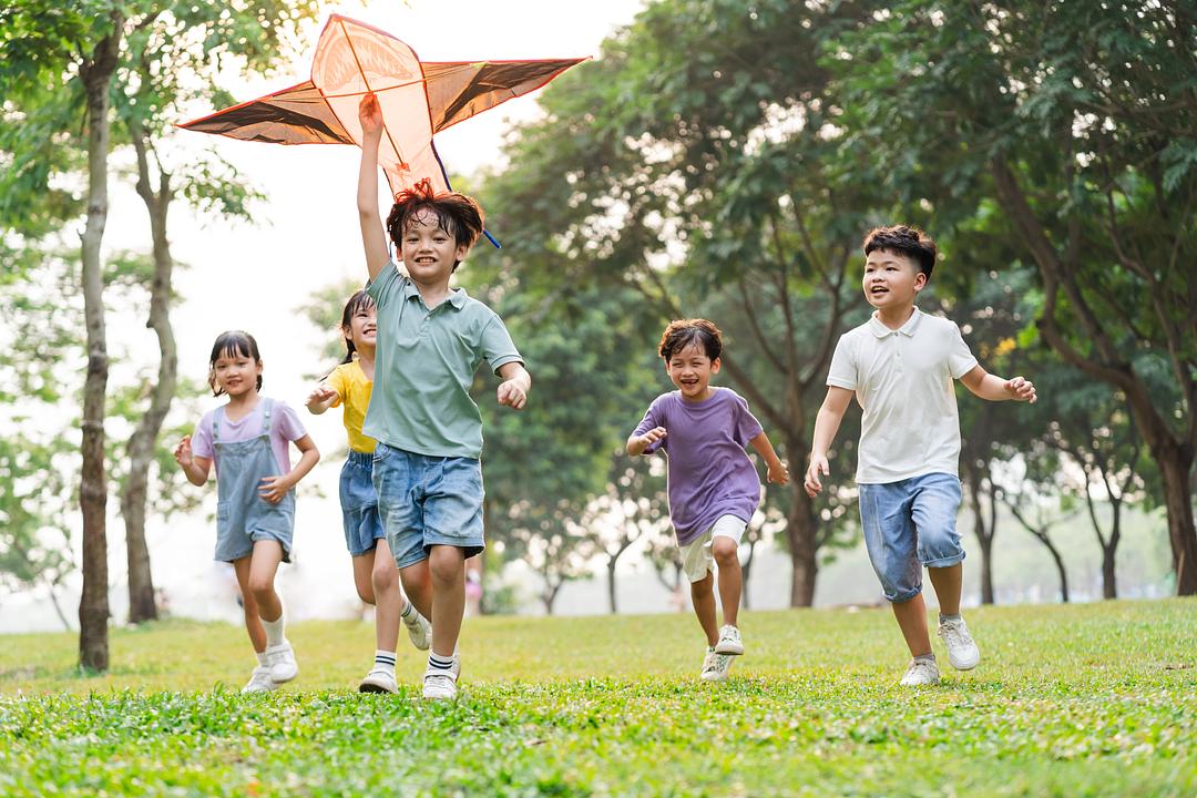 Group of children running through a park with a kite, modeled by children playing outdoors among trees and grass