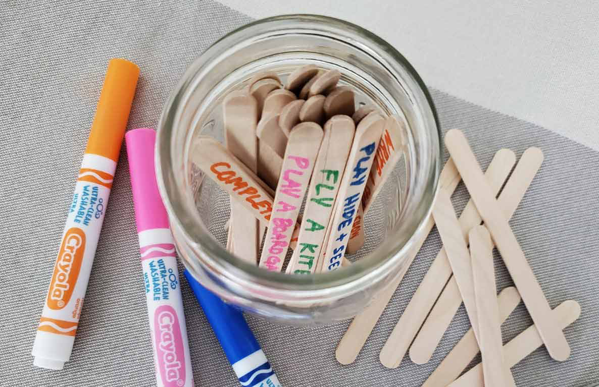 Mason jar filled with wooden craft sticks labeled with activity ideas, surrounded by Crayola markers and colored pencils on gray surface