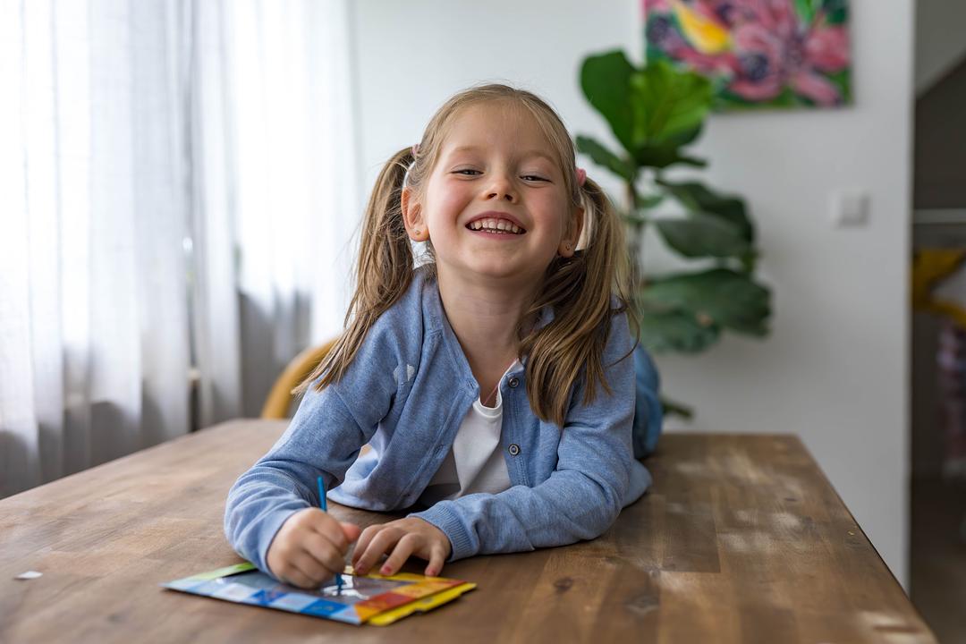 Young girl smiling while drawing with markers at a wooden table, modeled by a child in a blue shirt
