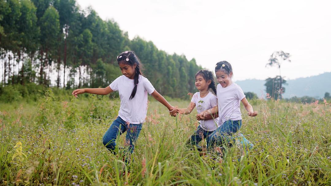 Three children running through a wildflower field with forested hills in the background, modeled by children in white shirts and jeans