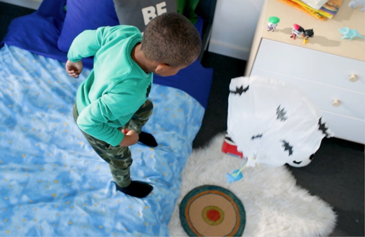 Child in green jacket playing parachute drop zone activity on blue mat, reaching toward target on floor in playroom setup