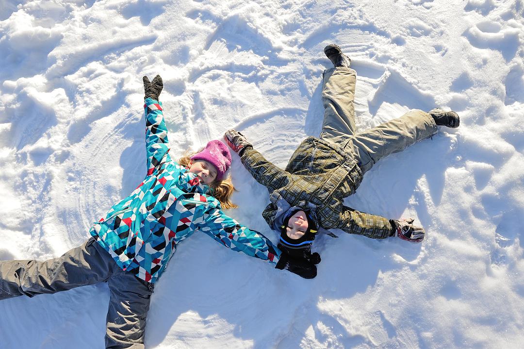 Three children lying in snow making snow angels, wearing winter coats and hats on a sunny day