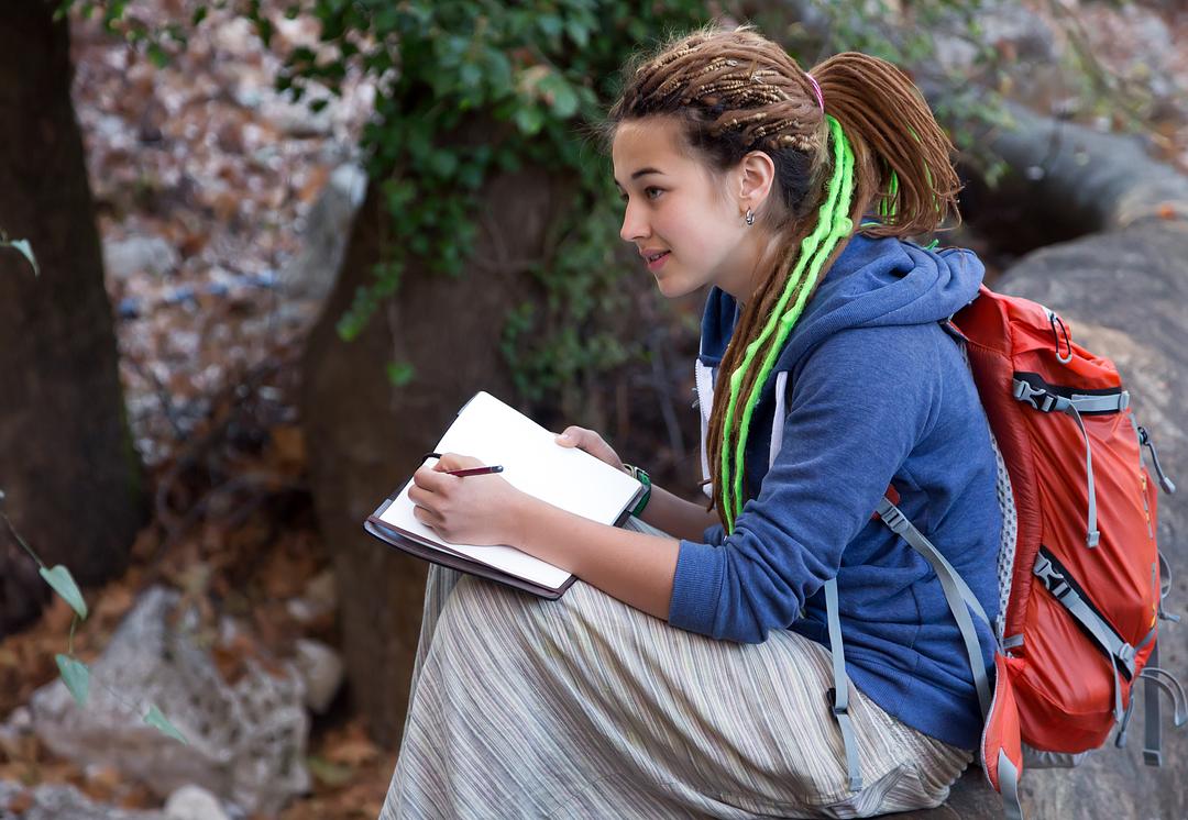 Child on model wearing blue hoodie and red backpack, holding clipboard and pen, standing outdoors against ivy-covered brick wall