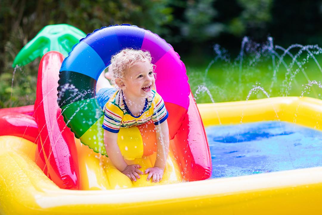 Toddler on model playing in colorful inflatable water play center with slide and sprinklers in backyard
