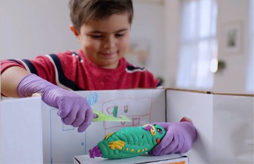 Child on model wearing purple gloves handling a green and yellow plush toy caterpillar over a cardboard box labeled with activity instructions