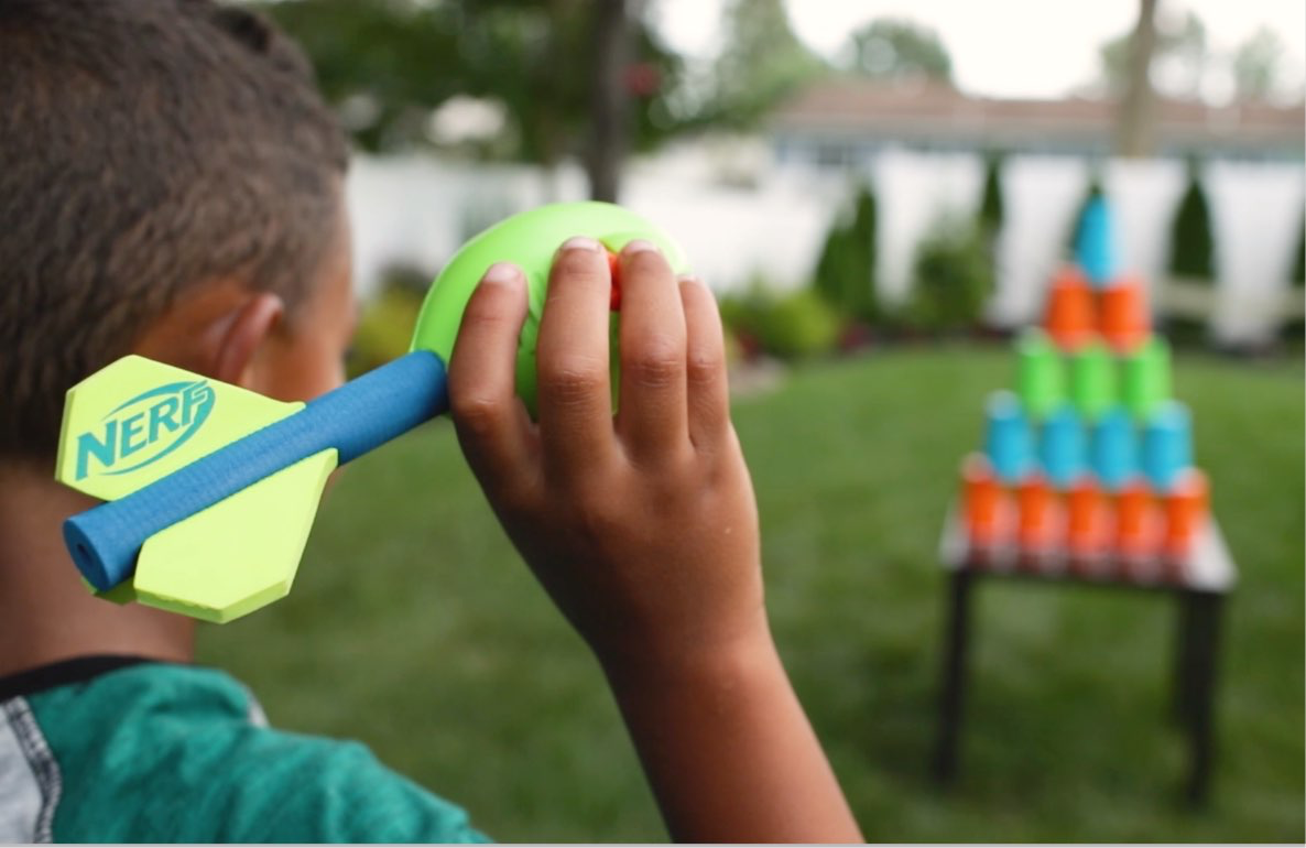 Child holding a Nerf blaster with foam dart, aiming at colorful stacked cups on a table in a backyard setting