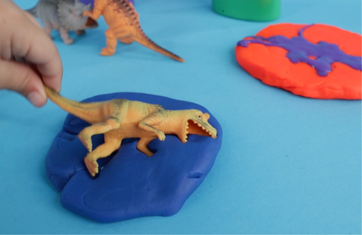 Child's hand placing a yellow dinosaur figure into a blue clay fossil mold, with additional dinosaur toys and colorful clay molds on a light blue surface
