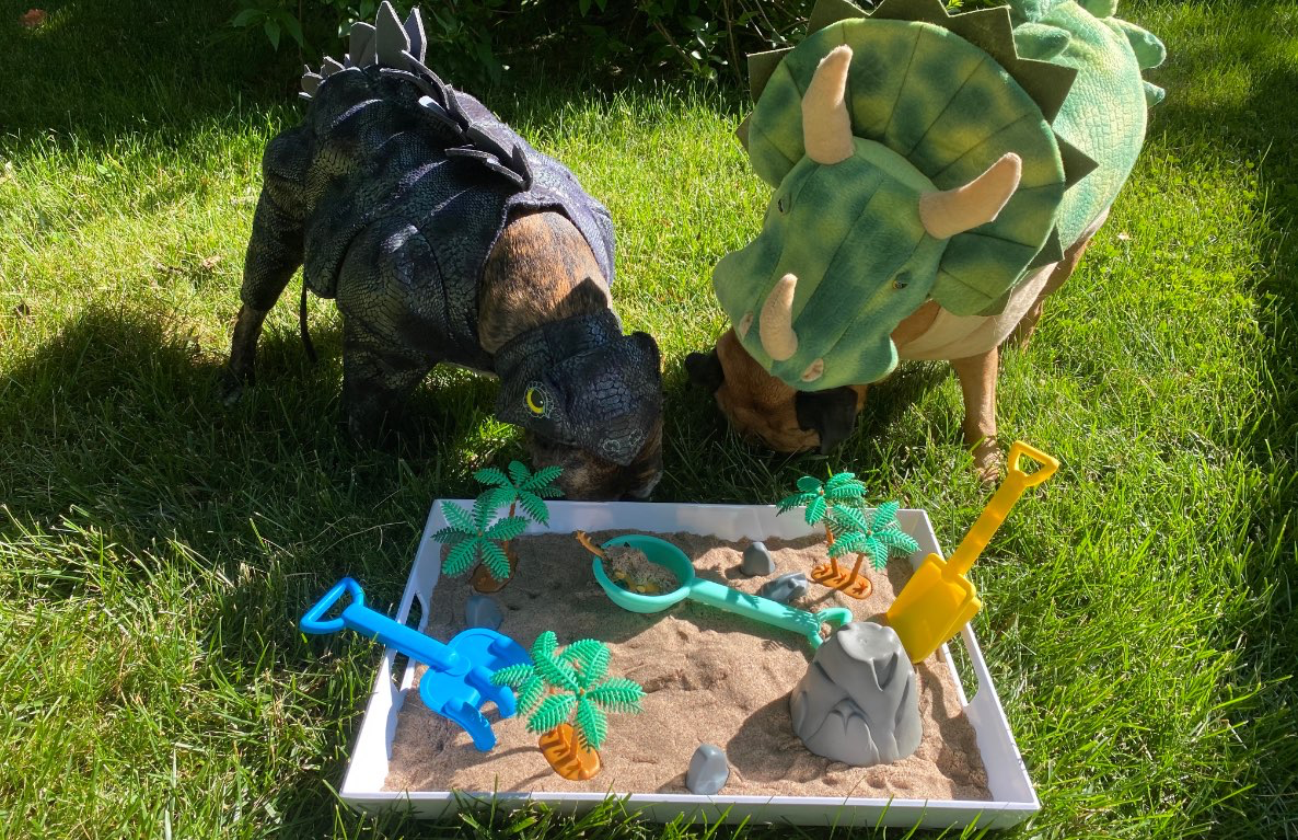 Two children wearing dinosaur costumes playing with a dinosaur-themed sandbox activity featuring toy dinosaurs, palm trees, a watering can, and excavation tools