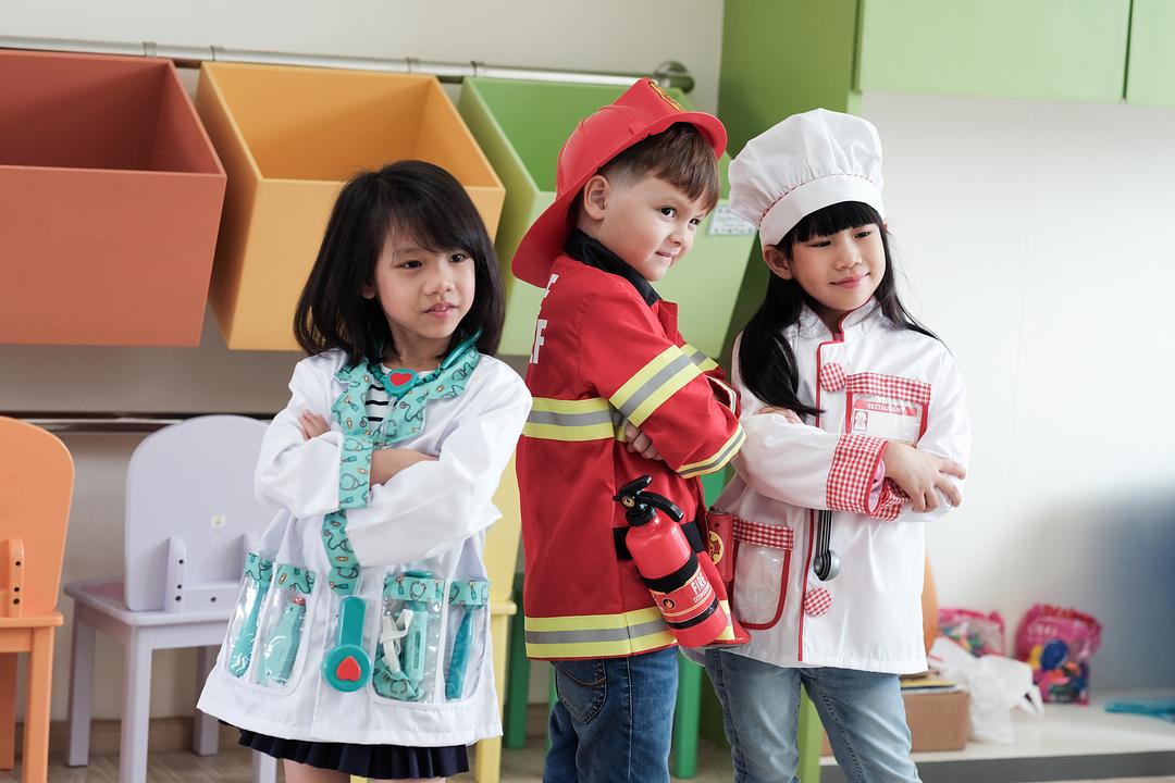 Three children modeled in pretend play costumes: doctor with stethoscope, firefighter with red jacket and cap, and chef with white hat and apron, in a colorful