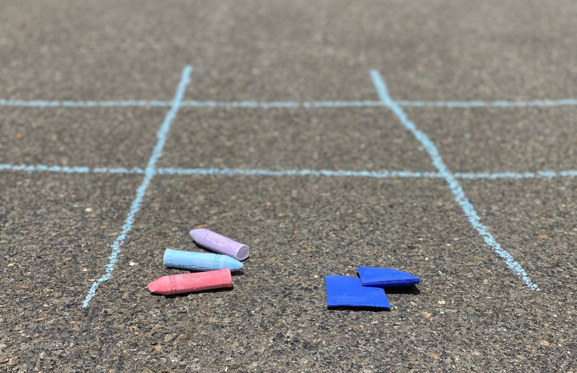 Chalk-drawn tic-tac-toe grid on asphalt with colored chalk pieces and bean bags arranged for gameplay