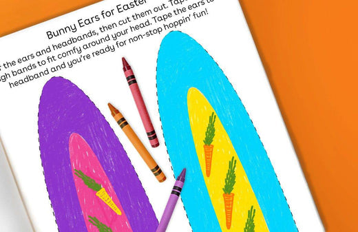 Colorful paper bunny ears craft project with purple and blue ear shapes decorated with yellow and green details, alongside crayons on an orange background
