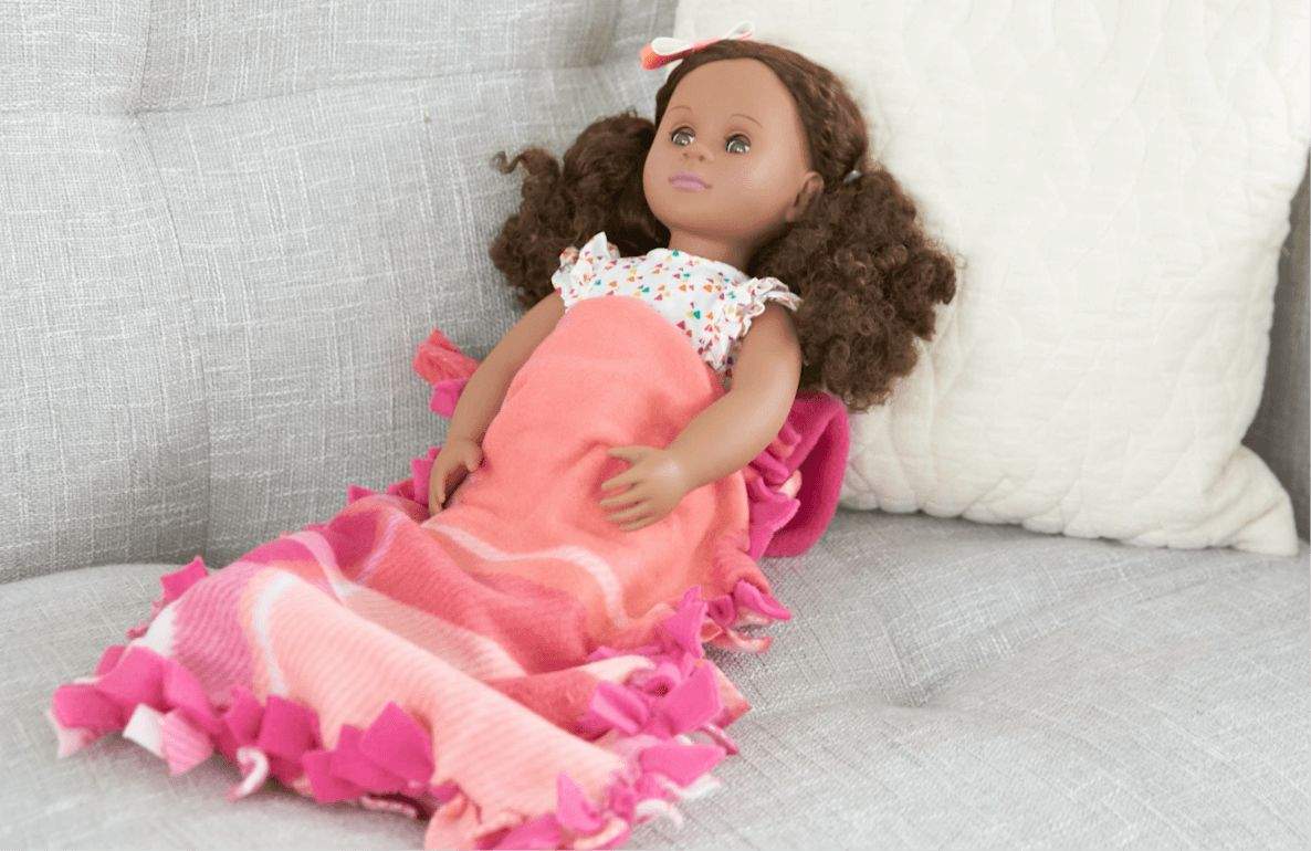 Doll with brown curly hair wearing a floral top and pink sleeping bag with ruffled edges, positioned on a gray couch