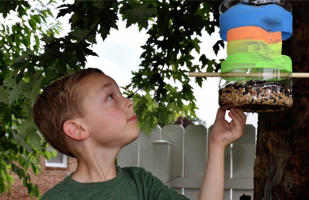 Child on model looking up at colorful stacked bird feeder hanging from tree branch in outdoor backyard setting