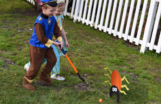 Two children playing DIY monster golf outdoors, one child swinging a blue club toward an orange felt monster decoration with googly eyes on grass, white fence i