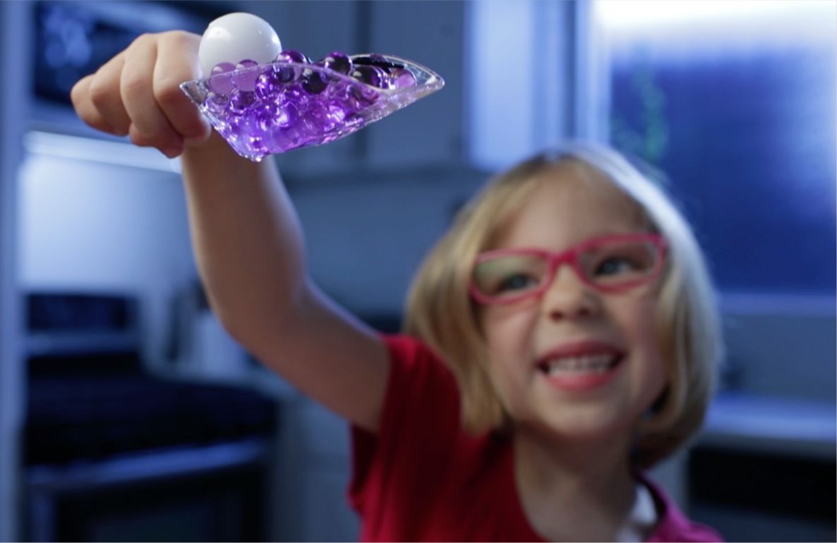 Child modeled by a girl holding purple glowing gel sensory toy, wearing pink glasses and red shirt