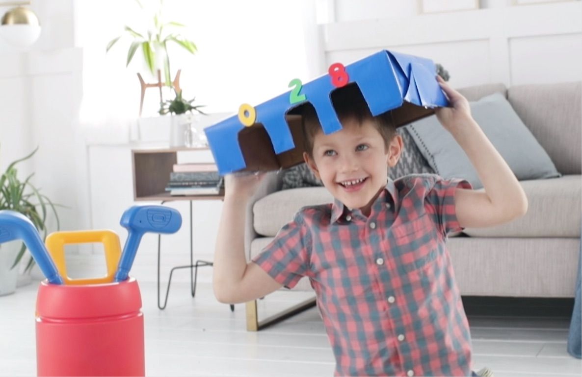 Child modeled by a child wearing red plaid shirt holding blue cardboard castle with numbered towers above head, with toy golf clubs in red bucket on living room