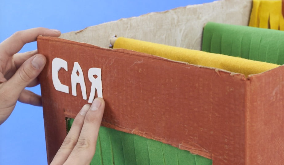 Child's hands holding a colorful cardboard box labeled "CAR" against a blue sky, part of a DIY Monster Jam car wash activity setup with yellow and green fabric