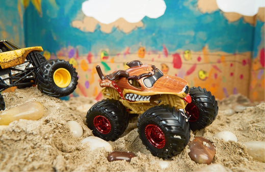 Monster truck toy with fierce mouth design on red wheels displayed in sand with colorful painted backdrop and another toy vehicle