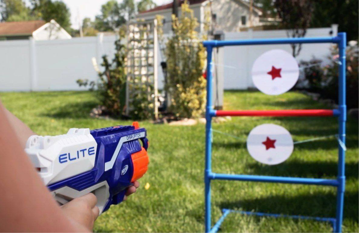 Child holding blue and white NERF Elite blaster, aiming at backyard target game with red stars on white circles mounted on blue frame
