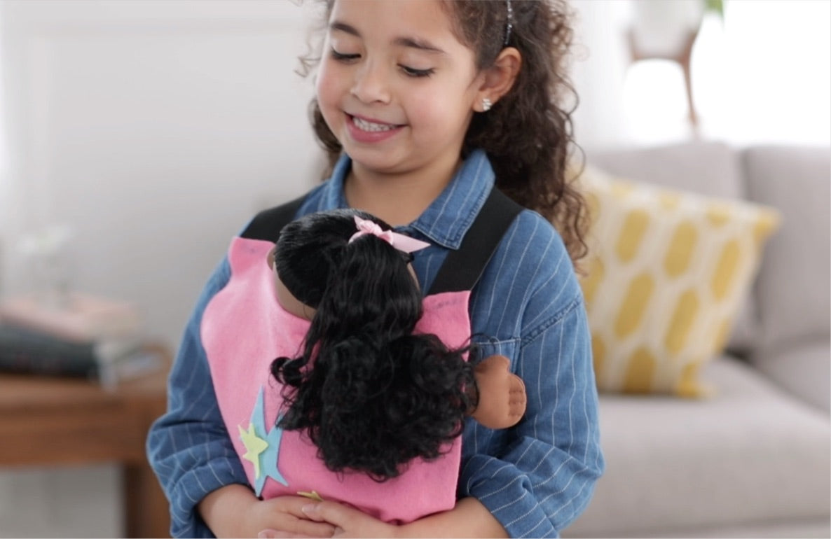 Child on model holding black plush doll with pink bow in homemade pink fabric carrier with colorful star design