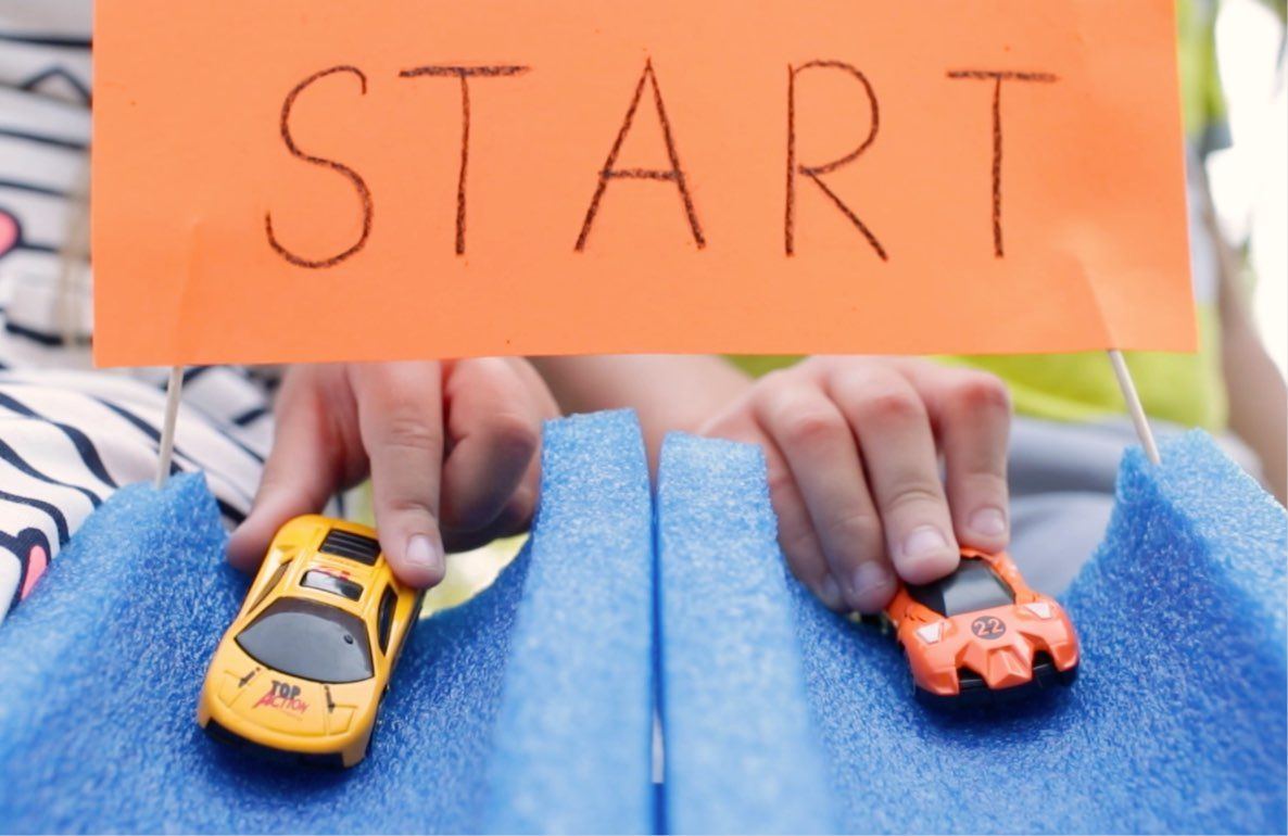 Child's hands holding a yellow toy car and orange toy car on blue pool noodles with orange sign reading START above