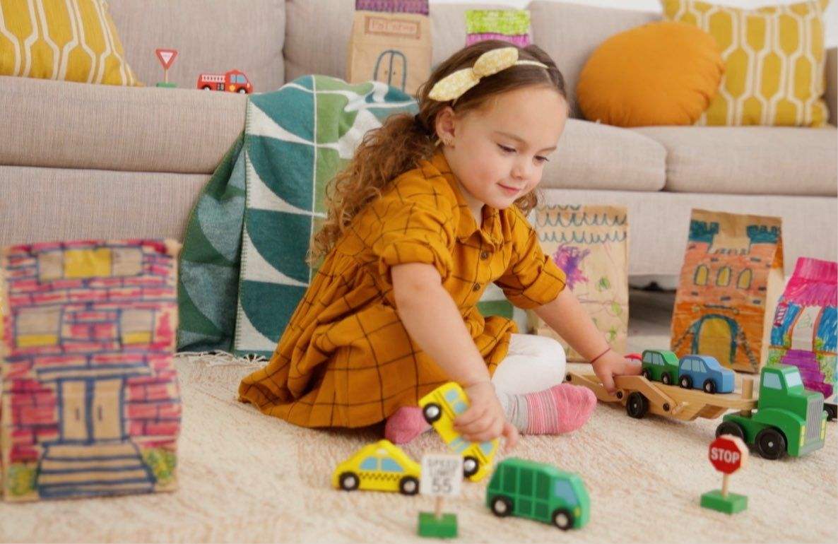 Child modeled by a girl playing with wooden toy vehicles and cardboard box buildings on a living room floor