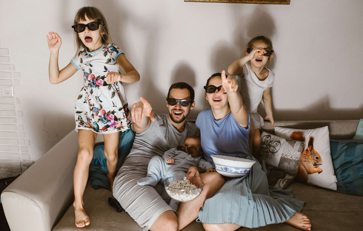 Family wearing sunglasses watching movies together on a couch, eating popcorn and snacks, laughing and celebrating