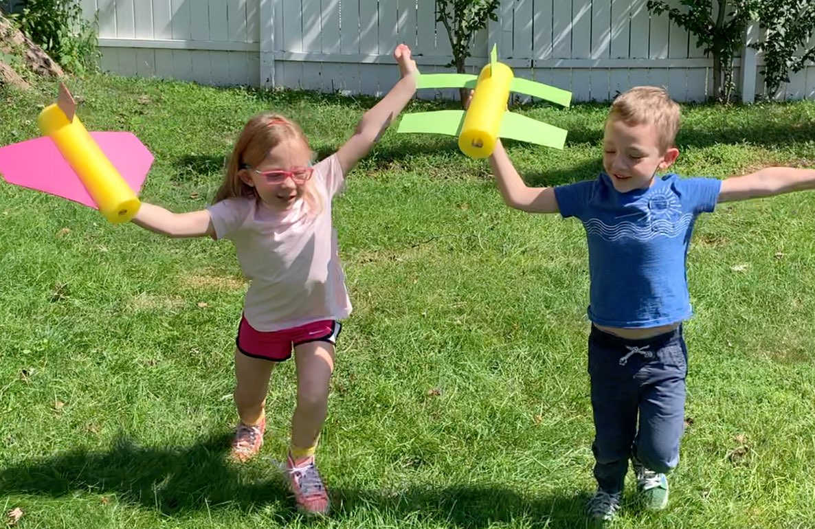 Two children playing with DIY pool noodle planes in a grassy outdoor space, modeled by a child on each side holding yellow and green foam constructions