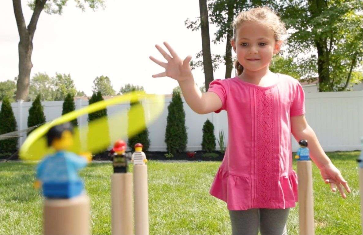 Child on model playing ring toss game outdoors on lawn with yellow rings and wooden posts, trees and white fence in background