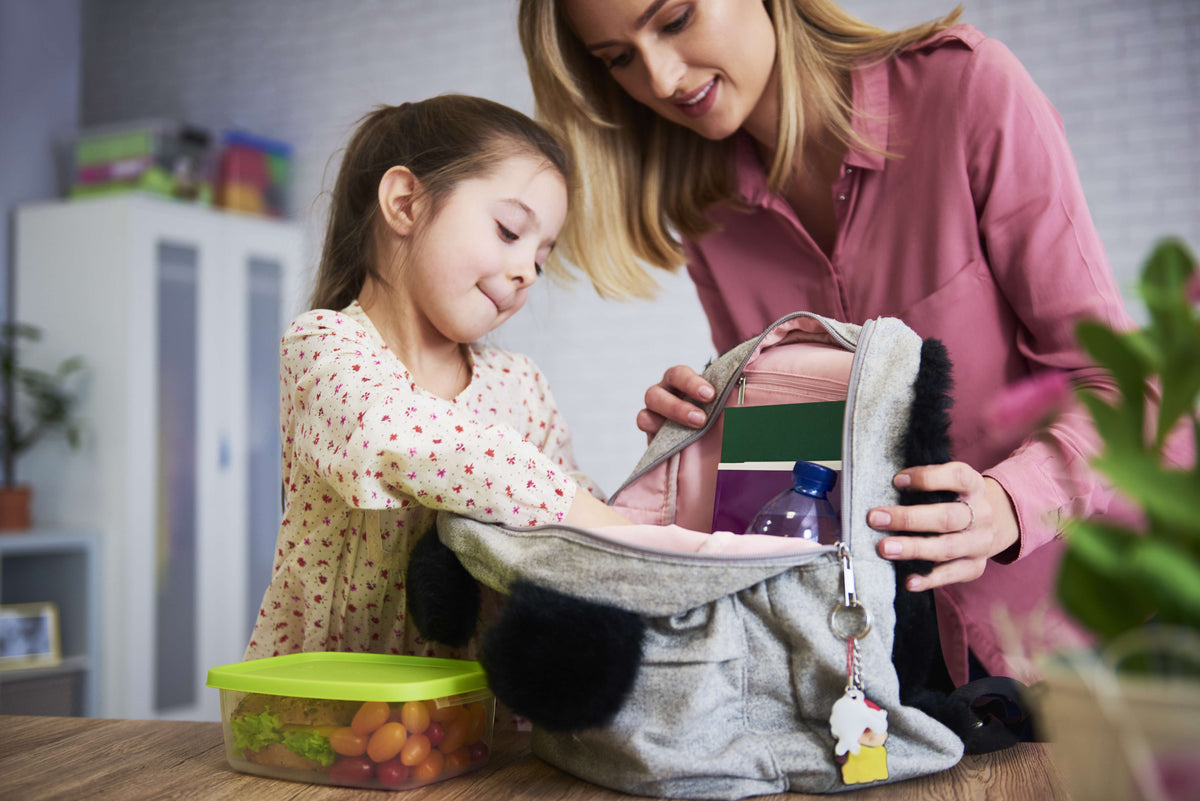 Adult and child opening a gray lunch bag with compartments, looking at colorful food containers inside on a kitchen table