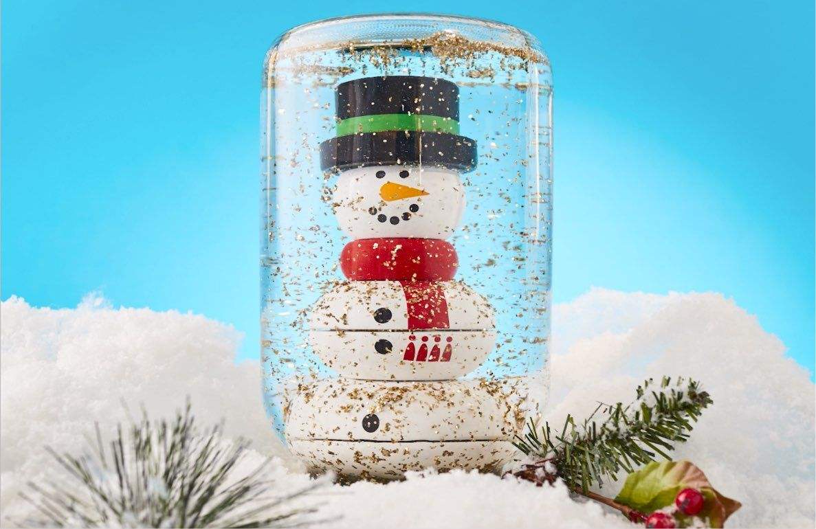 DIY snow globe craft with stacked wooden discs forming a snowman face, black top hat, green band, orange nose, coal eyes, red scarf, and black mouth details, su