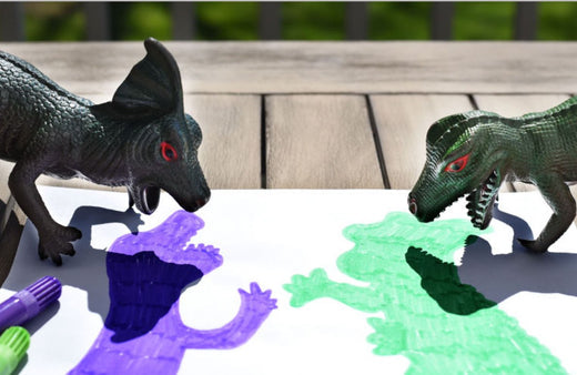 Two black dinosaur figures casting colorful shadows on white paper — purple shadow on left, green shadow on right — during a toy shadow tracing activity